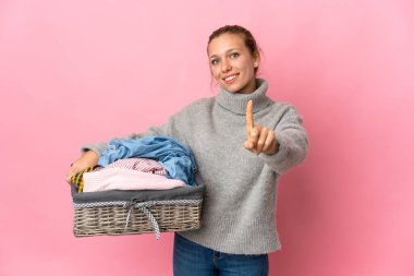 Pembe arka planda izole edilmiş çamaşır sepeti tutan genç bir kadın parmağını kaldırıyor ve gösteriyor.