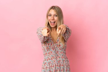 Pembe arka planda izole edilmiş genç sarışın kadın şaşırmış ve ön tarafı işaret ediyor.