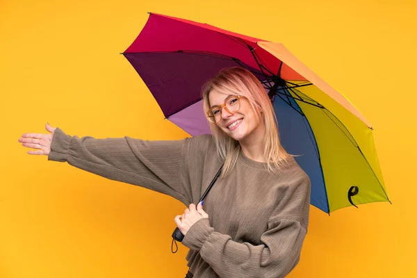 年轻的金发女子举着一把雨伞 越过孤立的黄墙 向旁边伸出手来 感谢她的邀请 — 图库照片