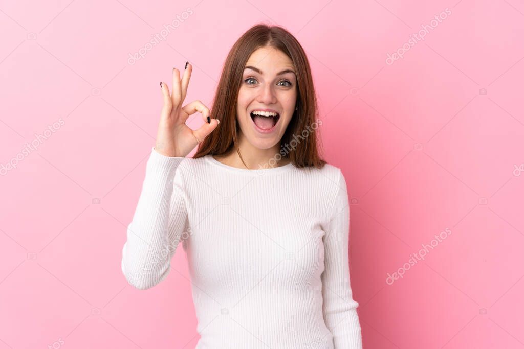 Young woman over isolated pink background surprised and showing ok sign