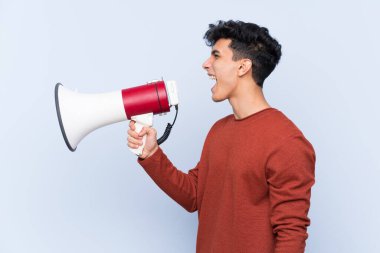 Arjantinli genç adam izole edilmiş mavi arka planda megafondan bağırıyor.