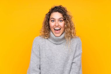 Sarışın, kıvırcık saçlı, balıkçı yaka kazak giyen, sarı arka planda, yüzünde sürpriz bir ifade olan genç bir kadın.