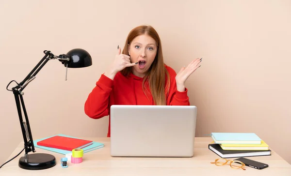 Young student woman in a workplace with a laptop making phone gesture and doubting