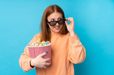 Üç boyutlu gözlüklü ve elinde koca bir kova patlamış mısırla izole edilmiş genç bir kızıl saçlı kadın.