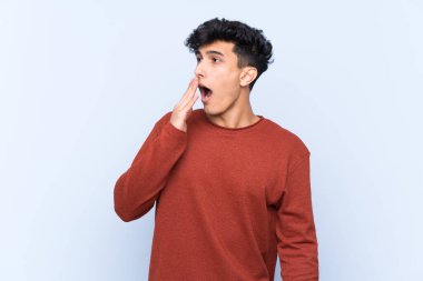 Arjantinli genç adam izole edilmiş mavi arka planda esniyor ve geniş bir ağzı eliyle kapatıyor.