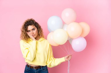 Kıvırcık saçlı genç bir kadın pembe arka planda izole edilmiş balonları yakalıyor mutsuz ve hayal kırıklığına uğramış.