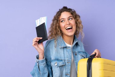 Kıvırcık saçlı genç bir kadın, mor arka planda izole edilmiş, bavul ve pasaportla tatilde.
