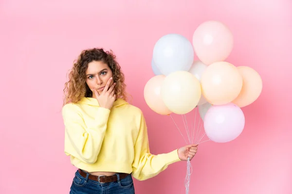 Young blonde woman with curly hair catching many balloons isolated on pink background thinking an idea