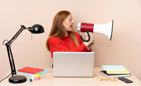 Young student woman in a workplace with a laptop shouting through a megaphone