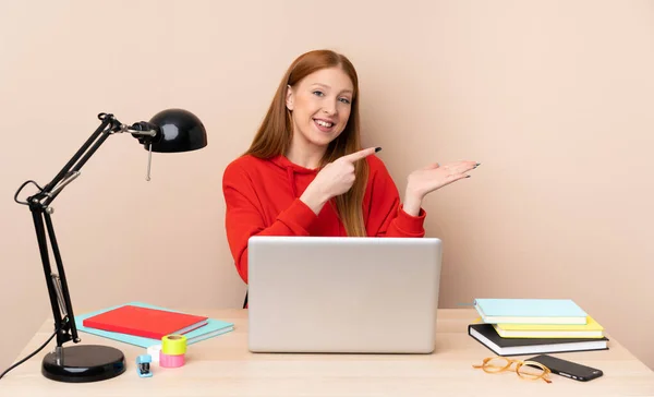 Young student woman in a workplace with a laptop holding copyspace imaginary on the palm to insert an ad