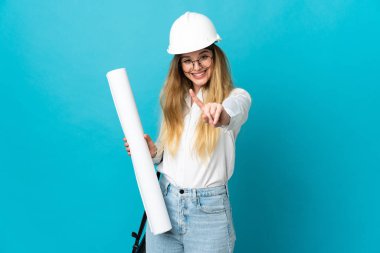 Miğferli ve elinde mavi arka planda izole edilmiş planları olan genç mimar kadın parmağını kaldırıyor.