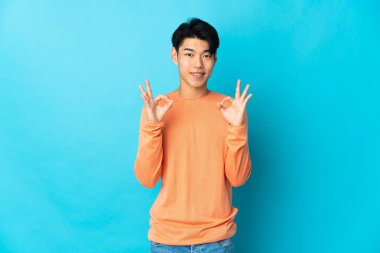 Mavi arka planda izole edilmiş genç bir Çinli iki eliyle tamam işareti gösteriyor.