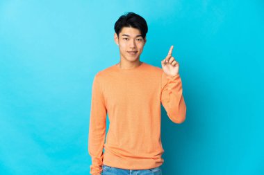 Genç Çinli bir adam mavi arka planda izole edilmiş ve en iyinin işareti olarak parmağını kaldırıyor.