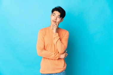 Genç Çinli bir adam mavi arka planda izole edilmiş, yukarı bakarken bir fikir düşünüyor.