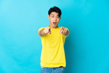 Mavi arka planda izole edilmiş genç bir Çinli şaşırmış ve ön tarafı işaret ediyor.