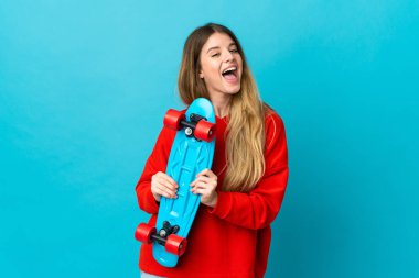 Mavi arka planda izole edilmiş genç sarışın kadın mutlu bir ifadeyle paten kayıyor.