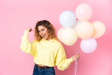 Kıvırcık saçlı genç bir kadın pembe arka planda izole edilmiş bir sürü balon yakalıyor. Güçlü bir jest yapıyor.