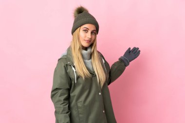 Pembe arka planda izole edilmiş, kış şapkalı genç bir kadın davet edilmek için ellerini yana uzatıyor.