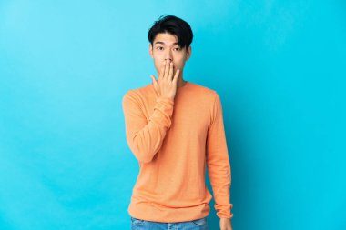 Genç Çinli adam mavi arka planda izole edilmiş. Ağzını eliyle kapatıyor.