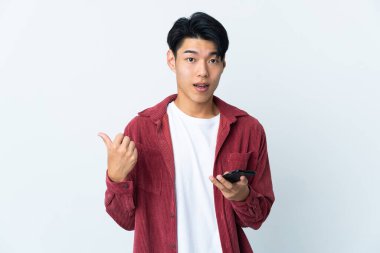 Genç Çinli adam beyaz arka planda cep telefonuyla izole edilmiş ve yan tarafı işaret ediyor.