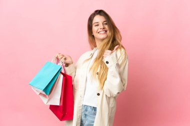 Alışveriş çantalı genç bir kadın pembe arka planda izole edilmiş. Elinde alışveriş torbaları ve başparmağıyla.