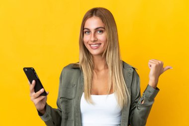 Sarı arka planda izole edilmiş genç sarışın kadın cep telefonu kullanıyor ve yan tarafı işaret ediyor.