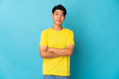 Genç Çinli adam mavi arka planda izole edilmiş. Kollarını ön cephede çapraz tutuyor.