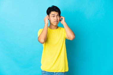 Mavi arka planda izole edilmiş genç bir Çinli hayal kırıklığına uğramış ve kulaklarını kapatmış.