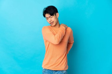 Genç Çinli bir adam mavi arka planda izole edilmiş, omuz ağrısı çekiyor, çabaladığı için.
