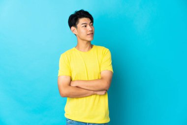 Genç Çinli adam mavi arka planda izole edilmiş kolları bağlı ve mutlu.
