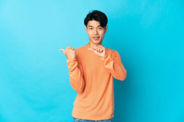 Mavi arka planda izole edilmiş genç Çinli bir adam bir ürün sunmak için kenarı işaret ediyor.