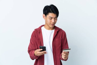 Genç Çinli bir adam beyaz arka planda izole edilmiş cep telefonuyla kredi kartıyla alışveriş yapıyor.