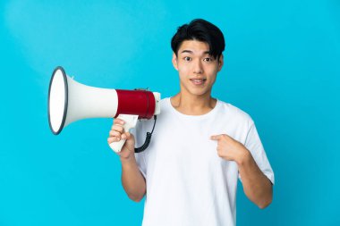 Genç Çinli bir adam mavi arka planda izole edilmiş elinde bir megafonla ve şaşırmış bir yüz ifadesiyle