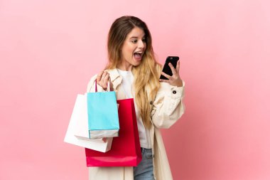 Alışveriş çantalı genç bir kadın pembe arka planda izole edilmiş alışveriş torbaları tutuyor ve bir arkadaşına cep telefonuyla mesaj yazıyor.