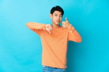 Genç Çinli adam mavi arka planda izole edilmiş, iyiye kötüye işaret yapıyor. Evet ya da hayır arasında kararsız