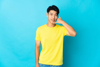 Genç Çinli bir adam mavi arka planda izole edilmiş bir şekilde telefon jesti yapıyor. Bana geri dönüş işareti ver.