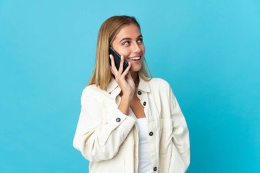 Mavi arka planda izole edilmiş genç sarışın kadın cep telefonuyla sohbet ediyor.