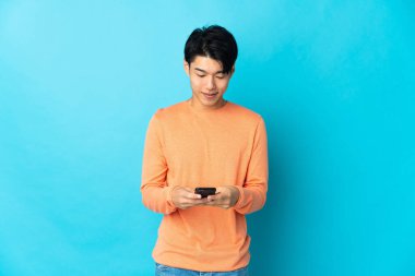 Genç Çinli adam mavi arka planda izole edilmiş cep telefonuyla bir mesaj gönderiyor.