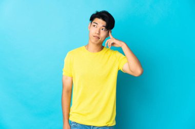 Genç Çinli bir adam mavi arka planda izole edilmiş. Delilik jesti yapıyor. Parmağını kafaya koyuyor.