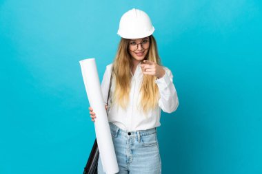 Kasklı ve elinde mavi arka planda izole edilmiş planları olan genç mimar kadın şaşırmış ve ön tarafı işaret ediyor.