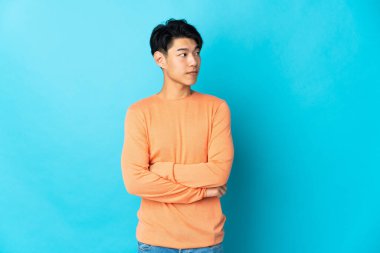 Genç Çinli adam mavi arka planda izole edilmiş, kollarını çapraz tutuyor.