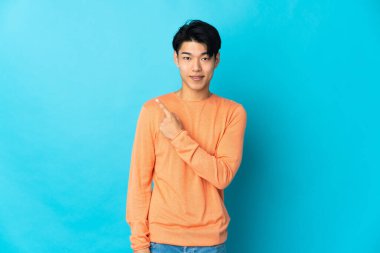 Mavi arka planda izole edilmiş genç Çinli bir adam bir ürün sunmak için kenarı işaret ediyor.