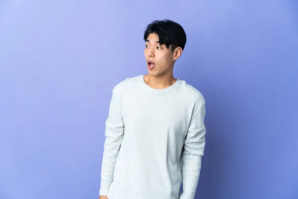 Young Chinese man isolated on purple background doing surprise gesture while looking to the side