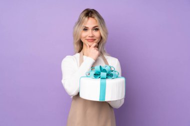 Genç pasta şefi mor arka planda büyük bir pasta tutuyor ve gülüyor.