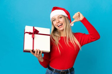 Noel şapkalı bir kadın mavi arka planda kendini beğenmiş ve gururlu bir hediye tutuyor.