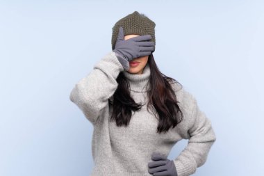 Kışlık şapkalı genç Kolombiyalı kız izole edilmiş mavi arka planda gözlerini elleriyle kapatıyor.
