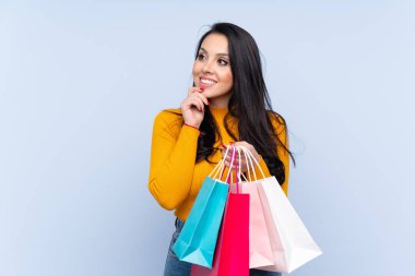 Genç Kolombiyalı kız izole edilmiş mavi arka planda elinde poşetlerle düşünüyor.