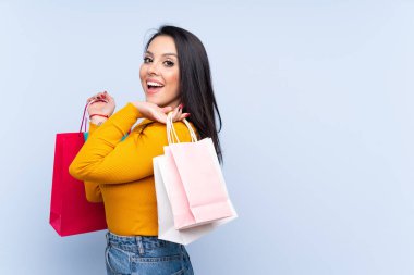 İzole edilmiş mavi arka planda genç bir Kolombiyalı kız alışveriş torbaları tutuyor ve gülümsüyor.