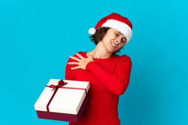 Noel şapkalı İngiliz kız, mavi arka planda izole edilmiş bir hediye tutuyor. Çabaladığı için omuz ağrısı çekiyor.