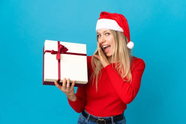 Noel şapkalı kadın elinde hediyeyle mavi arka planda izole edilmiş bir şekilde bir şeyler fısıldıyor.